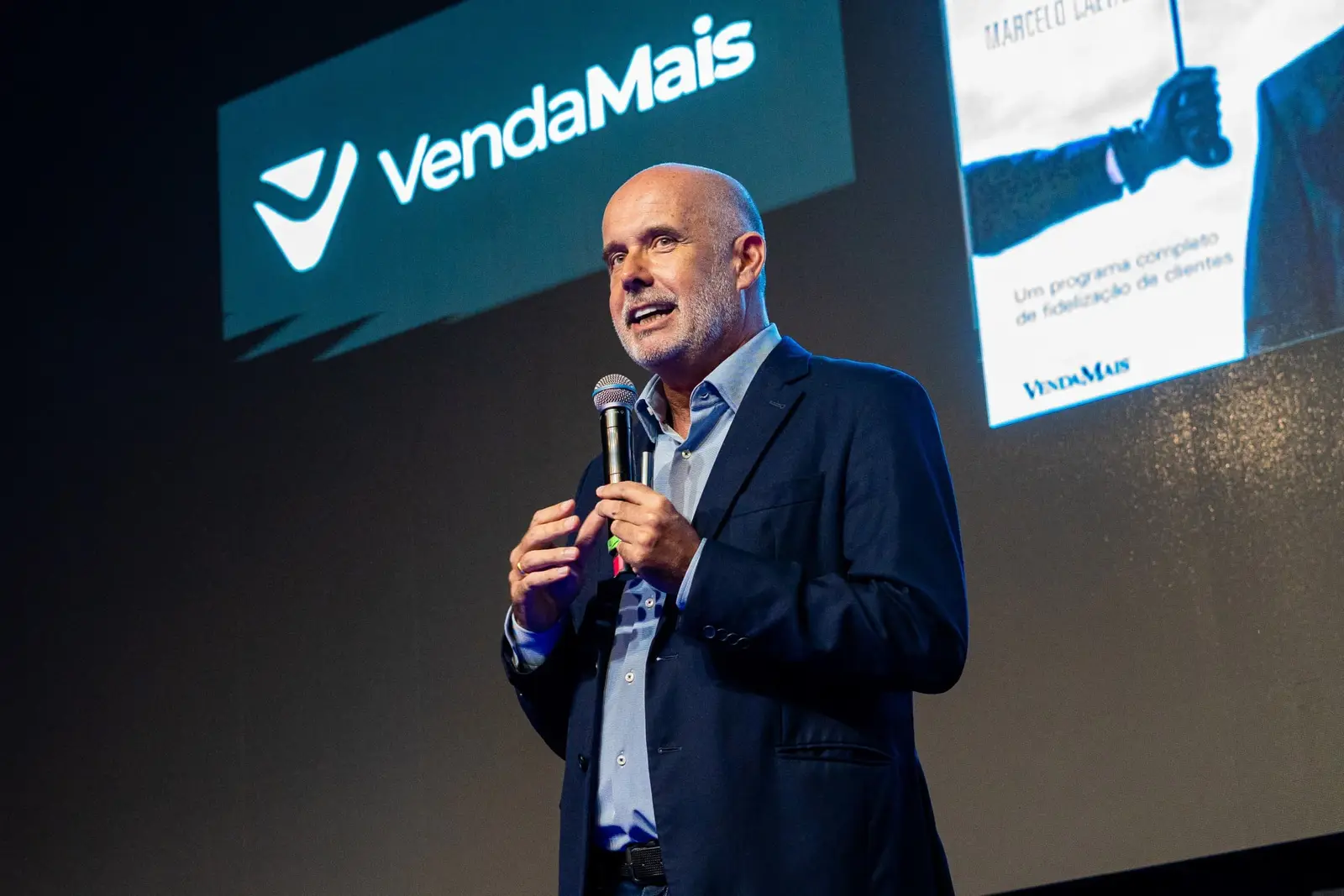 Marcelo Caetano falando ao microfone em um palco, com o logo da VendaMais projetado atrás